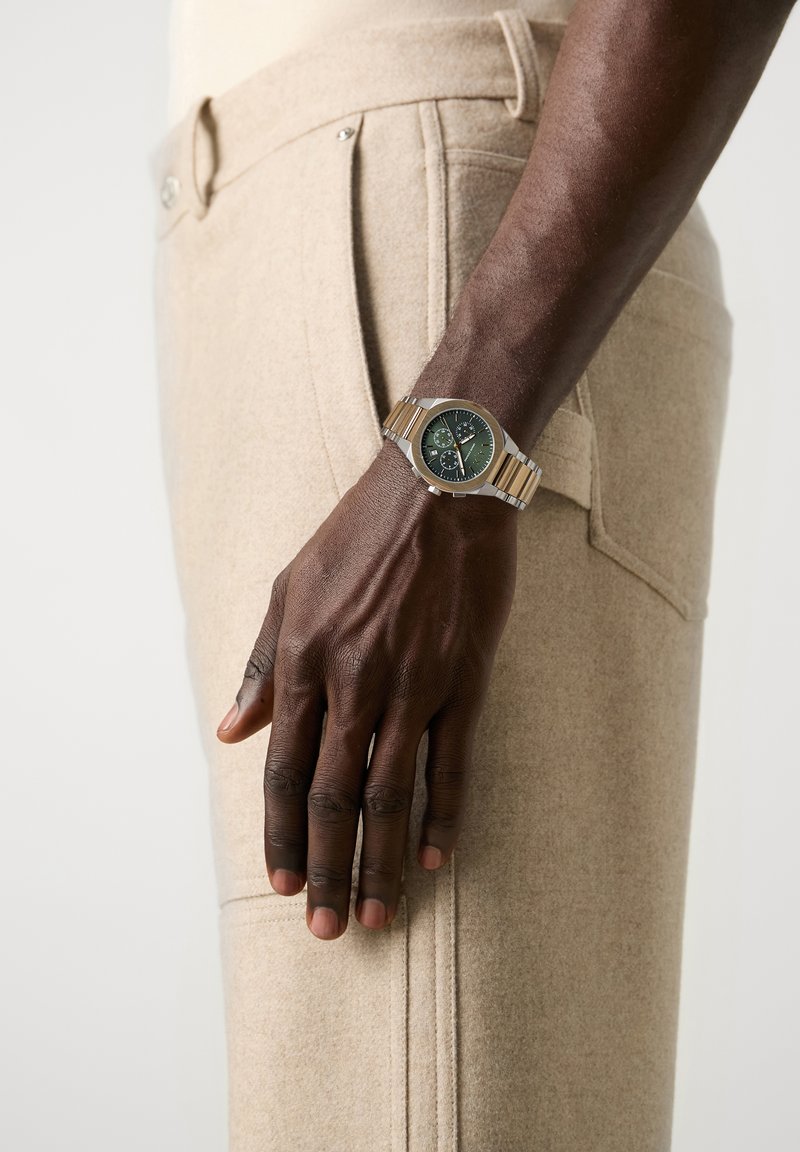 Dark-skinned arm wearing a silver and gold wristwatch with a green dial, resting beside beige pants with visible pocket stitching.