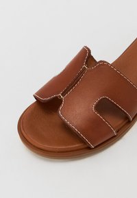 Brown leather sandal with two wide straps, exposed stitching, and a flat sole. Smooth texture and a rounded toe design.