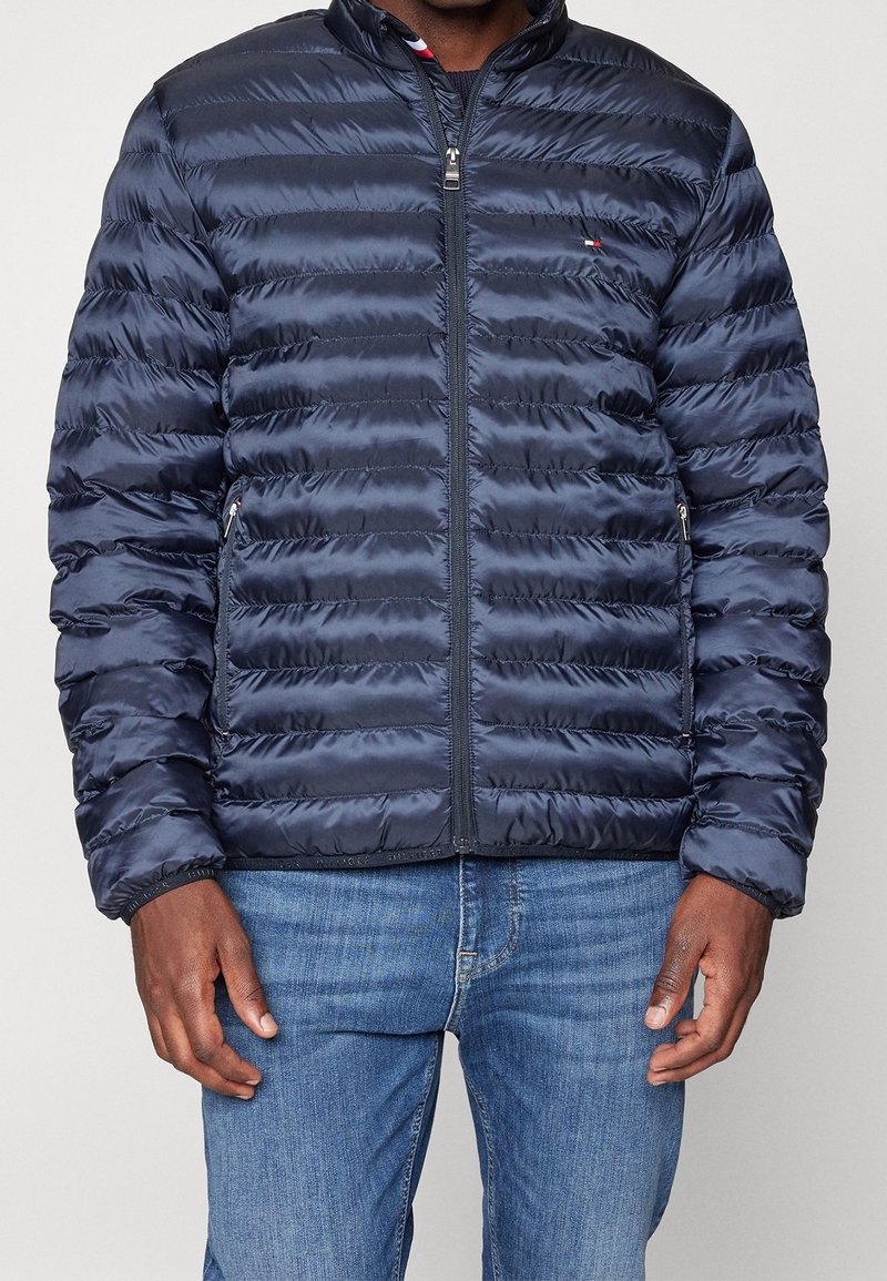 Granatowa kurtka puchowa z poziomymi szwami, wysokim kołnierzem, przednim zamkiem i dwiema bocznymi kieszeniami, wykonana z błyszczącego, lekkiego materiału.