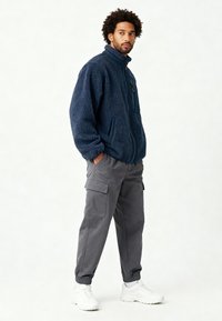 Veste polaire bleu marine avec col montant, fermeture éclair frontale et poche avant ; associée à un pantalon cargo gris et des baskets blanches.