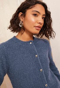 Tekstūru zeķubikses zils kardigans ar apaļu kakla izgriezumu, ar sudraba pogu akcentiem un pūkainu apdari. Mati veidoti brīvos cirtos.