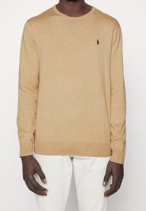 Mann trägt einen beigefarbenen Langarm-Pullover mit Rundhalsausschnitt und kleinem marineblauem Logo auf der Brust, kombiniert mit weißer Hose.