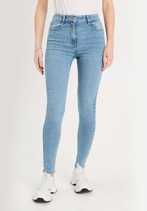 Kvinde iført lyseblå, højtaljede skinny jeans med fem lommer og hvide chunky sneakers, stående mod en ensfarvet baggrund.