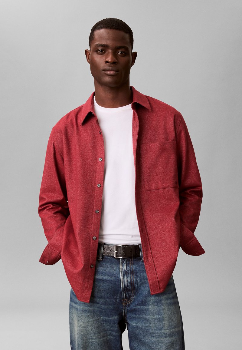 Chemise rouge boutonnée avec un motif texturé, portée sur un t-shirt blanc à col rond. Associée à un jean bleu et une ceinture foncée.