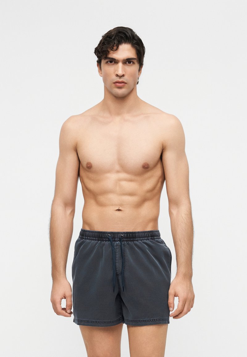 Joven con cabello corto y oscuro, vistiendo pantalones cortos de baño gris oscuro, de pie mirando hacia adelante contra un fondo claro y sencillo.