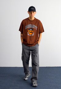Brązowy t-shirt graficzny z logo "Carhartt", szare spodnie cargo z kieszeniami i czapka w kolorze czarnym. Nosi w połączeniu z szaro-żółtymi trampkami. Proste tło.