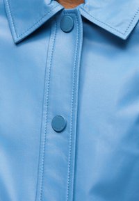 Φωτεινό γαλάζιο δερμάτινο πουκάμισο με δύο κουμπιά πάτημα και δομημένο γιακά που παρουσιάζει λεπτομερή ραφή.