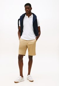 Bela polo majica, bež kratke hlače, temno modra pulover vržena čez ramena, bele superge, stoječi položaj na enostavni podlagi.