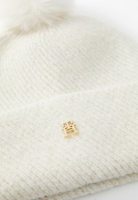 Valge kootud beanie pehme pom-pomiga üleval, millel on kuldne logoaksent ja peen sära kogu kangal. Tekstuur näeb välja pehme.