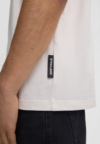 Korte mouw beige T-shirt met een rechte snit, voorzien van een zwart label gestikt op de zijnaad. De stof lijkt zacht en glad te zijn.