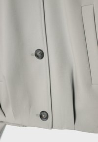 Section de manteau en tissu beige clair avec deux boutons ronds foncés et une poche passepoilée verticale sur le côté droit.