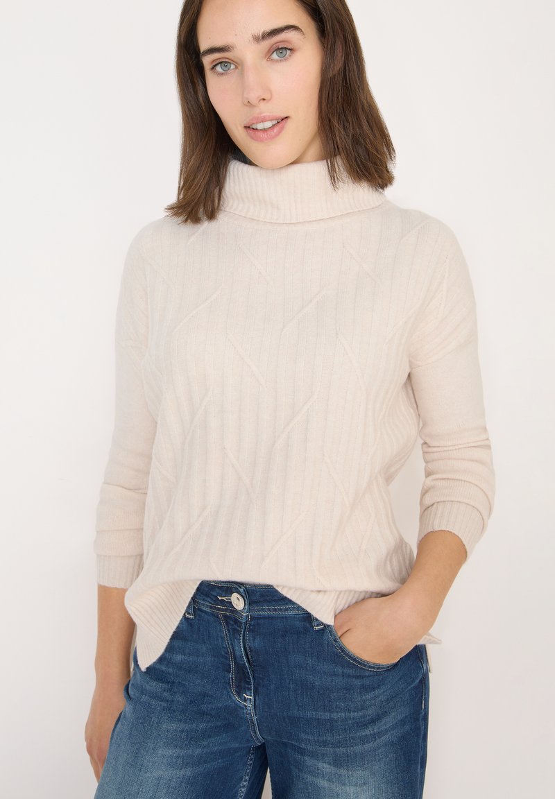 Lys beige turtleneck sweater med ribbet tekstur og diagonal mønster, parret med blå jeans. Sweateren har lange ærmer og slides i siderne.