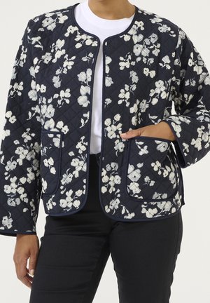 Femme portant une veste matelassée bleu marine avec un imprimé floral blanc, des poches avant, sans col, sur un t-shirt blanc et un pantalon noir.