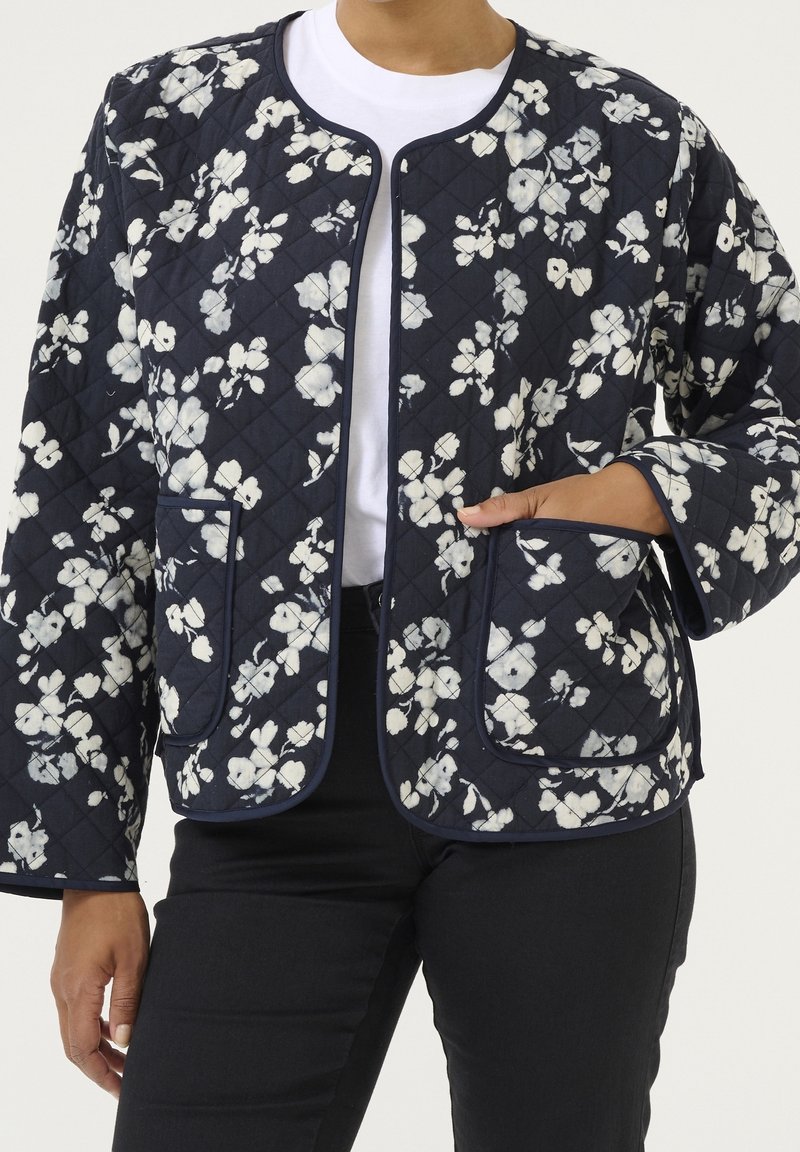 Femme portant une veste matelassée bleu marine avec un imprimé floral blanc, des poches avant, sans col, sur un t-shirt blanc et un pantalon noir.