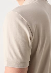 Gros plan sur l’épaule et le haut du bras d’une personne portant une chemise à manches courtes en tissu texturé beige clair.
