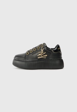 Black leather sneaker with a chunky ridged sole, gold accents, decorative lace hardware, and a branded logo detail on the side.