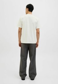Tricou din bumbac de culoare bej deschis pentru bărbați, cu mâneci scurte și decolteu rotund, purtat cu pantaloni largi de culoare gri închis și pantofi slip-on negri.