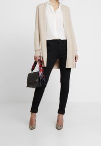 Beige long cardigan over a white shirt, black skinny jeans, and snake-print heels. Black bag with a colorful scarf detail.