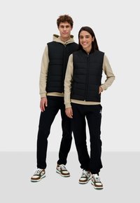 Czarne ocieplane kamizelki zapinane na zamek, noszone na beżowych bluzach z kapturem, uzupełnione granatowymi spodniami i brązowymi sneakersami.