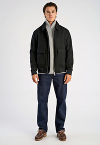 Svart jacka med dragkedja över en grå tröja med hög krage, mörkblå jeans och bruna skor. Jackan har två framfickor.