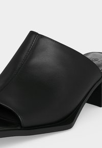 Μαύρο δερμάτινο mule με ανοιχτή μύτη, χαμηλό τετράγωνο τακούνι και λείο, άψογα ραμμένο επάνω μέρος που καλύπτει το μπροστινό μέρος του ποδιού.