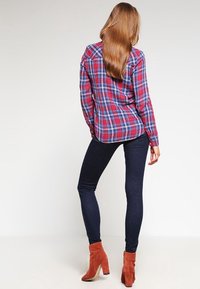 Vrouw met lang haar in een rood-blauw geruit overhemd, donkere skinny jeans en roestkleurige enkellaarsjes met hakken, staand tegen een effen achtergrond.