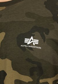 Roheline kamuflaažiga T-särk, mis on valmistatud puuvillast, lühikeste varrukatega, ümmarguse kaelusega ning esiküljel on valge "Alpha Industries" logo.
