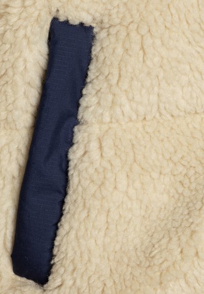 Beige gyapjú textúra, puha, bolyhos felülettel; jellemzője egy sötétkék szövetpanel sima felülettel. Látható varrások a szélek mentén.