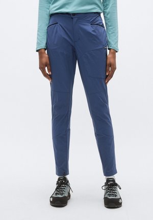 Kék, könnyű anyagból készült kültéri nadrág, combján zsebekkel, szűkített fazonú, megerősített varratokkal. Fekete túracipővel viselve.