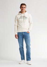 Helle beige Kapuzenjacke mit grünem Aufdruck, Kordelzugverschluss und gerippten Bündchen, kombiniert mit blauen Jeans und weißen Turnschuhen.