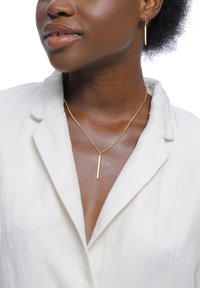 Smykkesæt i guld-tone med en slank bar-halskæde og matchende øreringe. Modellen bærer en lysefarvet skjorte med krave.