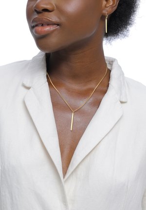 Smykkesæt i guld-tone med en slank bar-halskæde og matchende øreringe. Modellen bærer en lysefarvet skjorte med krave.