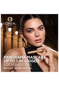 Woman with smooth hair and gold earrings holds gold tube of Panorama Mascara near her face, promoting bigger-looking eyes at night.