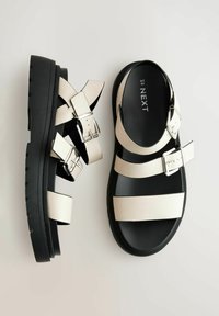 Sandali platform nere con cinturini crema, caratterizzati da dettagli con fibbia e una texture liscia. La suola presenta un motivo antiscivolo per una migliore aderenza.