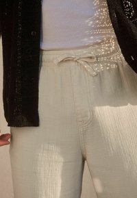 Pantalon beige clair en tissu texturé, doté d'une taille élastique et d'un cordon de serrage. Une chemise à motifs noire se superpose.