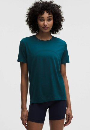 Vrouw met krullend haar, die een turquoise T-shirt met korte mouwen en zwarte aansluitende korte broek draagt, staand tegen een effen witte achtergrond.