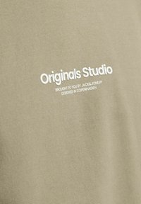Camiseta de color verde oliva claro con tejido suave y texturizado. Presenta texto impreso en blanco: "Originals Studio", "brought to you by JACK&JONES" y "diseñado en Copenhague."