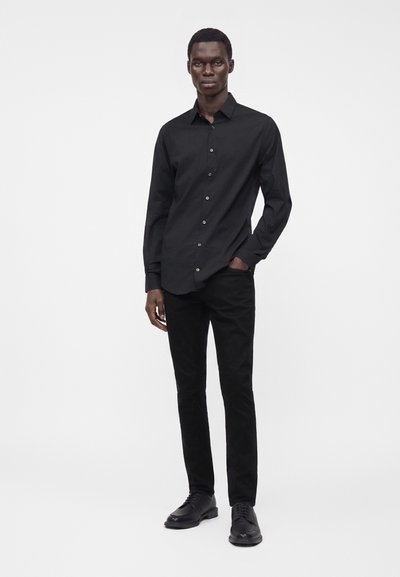 Uomo in piedi che indossa una camicia nera attillata con bottoni, pantaloni neri e scarpe eleganti nere su uno sfondo chiaro e semplice.