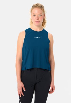 Junge Frau mit lockigem blondem Haar trägt ein blaues ärmelloses Oberteil und schwarze Hose, steht vor einem schlichten weißen Hintergrund.