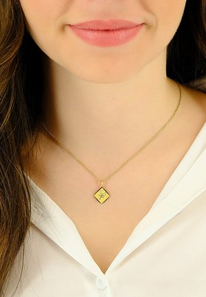 Collier pendentif en or avec un charm en forme de diamant présentant des détails complexes, suspendu à une fine chaîne, porté sur une blouse blanche.