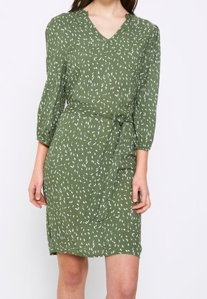 Kobieta ubrana w zieloną sukienkę o długości do kolan z białym, abstrakcyjnym wzorem, z dekoltem w serek, rękawami 3/4 oraz zawiązanym paskiem w talii na białym tle.