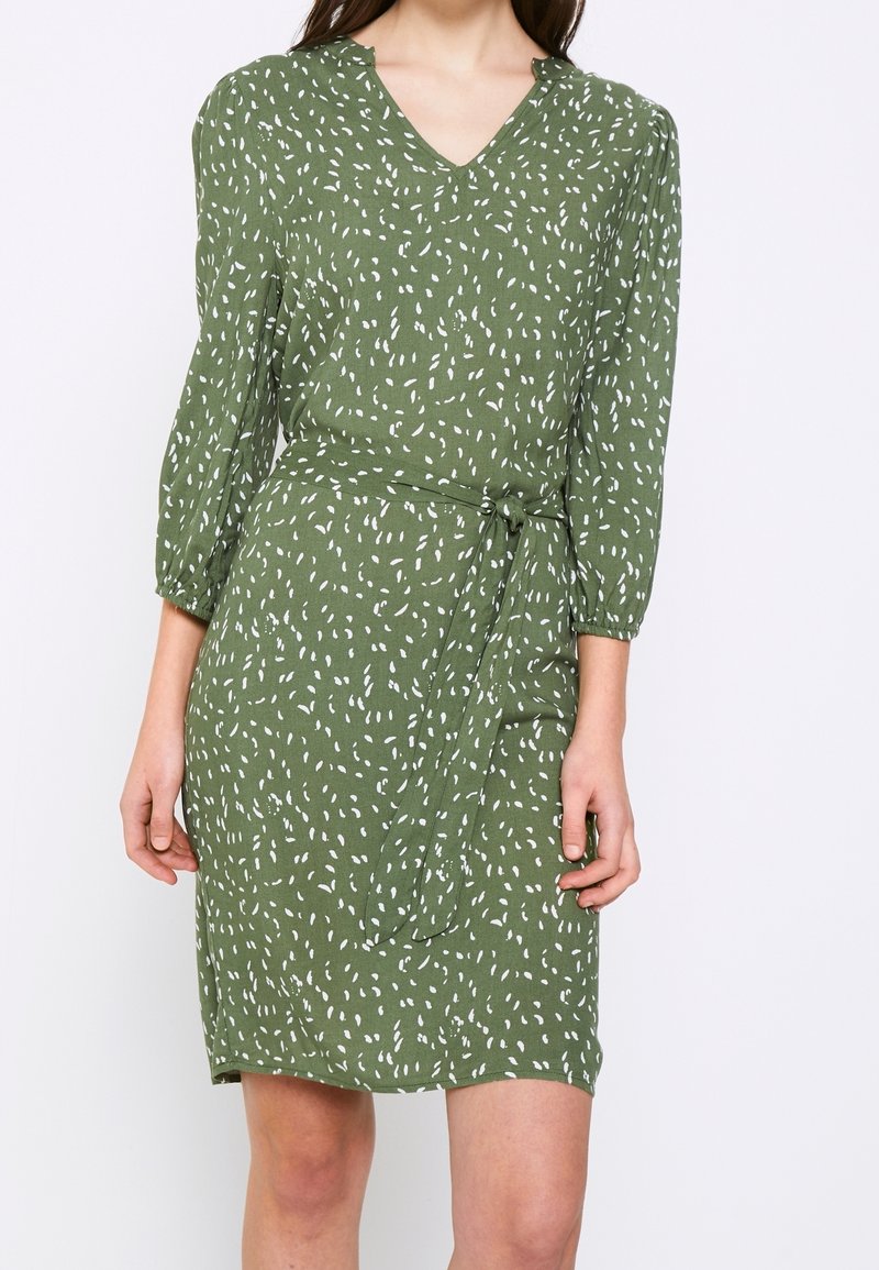 Femme portant une robe verte mi-longue avec un imprimé abstrait blanc, col en V, manches trois-quarts et ceinture nouée à la taille, sur fond blanc.