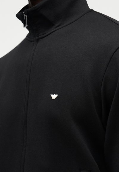 Juoda užtraukiama striukė iš minkštos medžiagos, turinti aukštą apykaklę ir mažą baltą siuvinėtą logotipą kairėje krūtinės srityje.