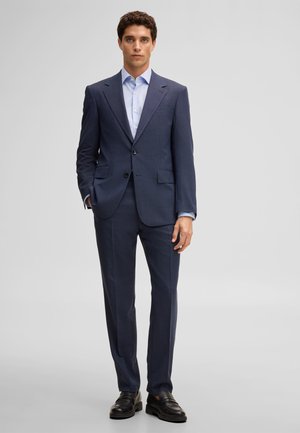 Mann steht aufrecht, trägt einen dunkelblauen Anzug mit hellblauem Hemd und schwarzen Anzugsschuhen vor einem einfarbigen Hintergrund.