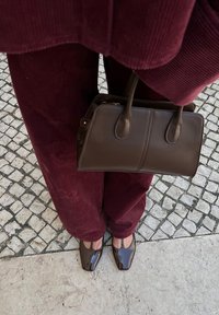 Borsa a tracolla in pelle marrone con due manici corti, abbinata a pantaloni in corduroy bordeaux e scarpe lucide marroni, il tutto su una superficie in ciottoli.