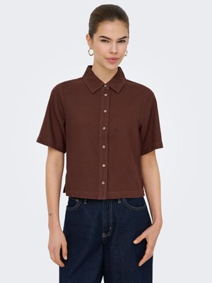 Sieviete valkā brūnu īsroku pogājamu krekli ar apkakli un tumši zilu augstas jostasvietas džinsu bikses, stāvot pret vienkāršu fonu.