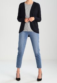 Navy cardigan dengan bagian depannya terbuka, kaos leher bulat abu-abu di bawahnya, dan jeans biru muda. Sepatu hak tinggi hitam menyelesaikan penampilan.