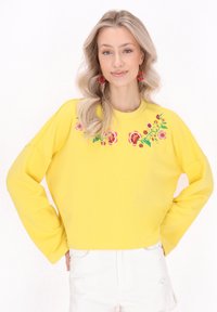 Felpa gialla con ricami floreali in rosa e verde, con vestibilità ampia e maniche larghe, abbinata a shorts di jeans bianchi.