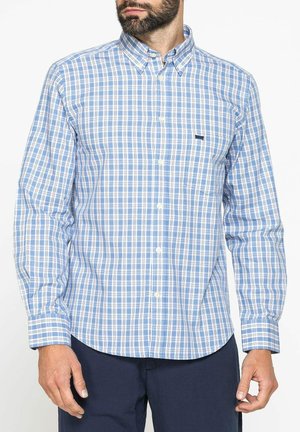 Uomo che indossa una camicia a quadri azzurri e bianchi con taschino frontale e pantaloni blu scuro, su sfondo semplice.