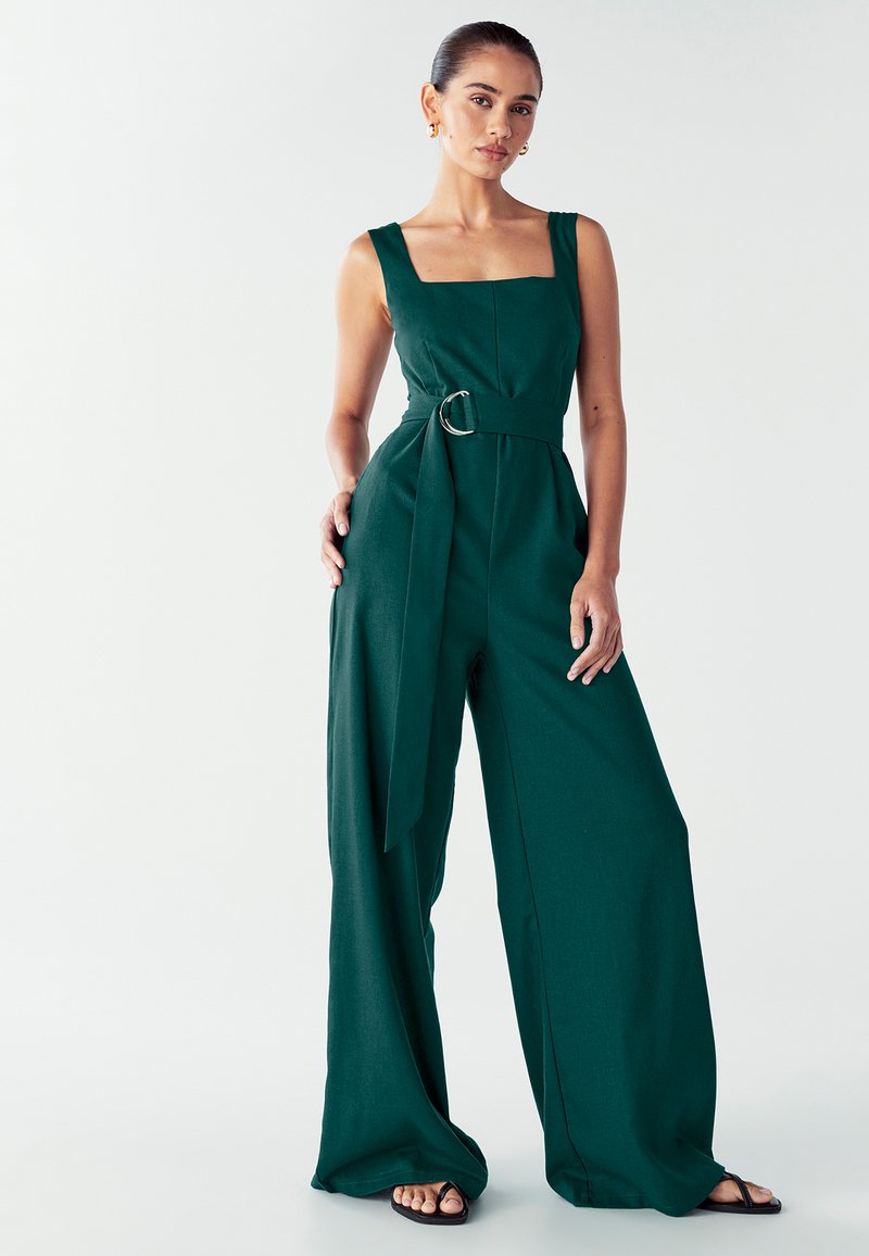 Mujer vestida con un mono sin mangas de color verde oscuro, ceñido con cinturón y de piernas anchas, de pie con una mano en el bolsillo y sandalias negras sobre fondo blanco.